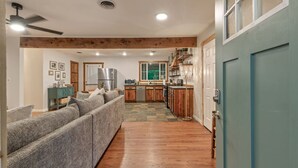 A spacious living room with a sofa, wooden flooring, and a kitchen area with wooden cabinets and a refrigerator. - Rustic Charm Owasso Retreat | 2BR • Big Backyard (Owasso)