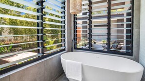 A modern bathroom with a freestanding bathtub, large windows with horizontal blinds, and a view of greenery outside.