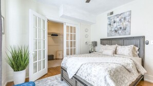 Une chambre à coucher avec un lit, une plante en pot, un banc bleu et une armoire dont les portes sont ouvertes.