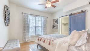 Une chambre à coucher comprenant un lit, un banc, un ventilateur de plafond, une fenêtre avec des rideaux et une armoire.