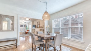 Un coin repas comprenant une table en bois, quatre chaises, un luminaire suspendu et une grande fenêtre avec des stores.
