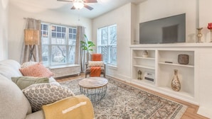 Un salon comprenant un canapé, un fauteuil, une table basse ronde et un téléviseur à écran plat fixé à un mur blanc.