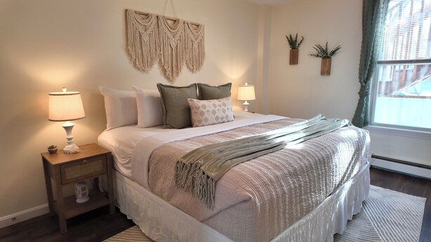 Un dormitorio ordenado con cama, lámparas de noche, un colgador de macramé y dos plantas en macetas.