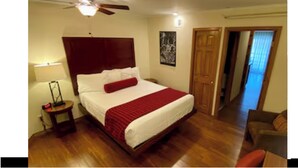 Een hotelkamer met een groot bed, een plafondventilator, een nachtkastje en een deur die naar een andere kamer leidt.