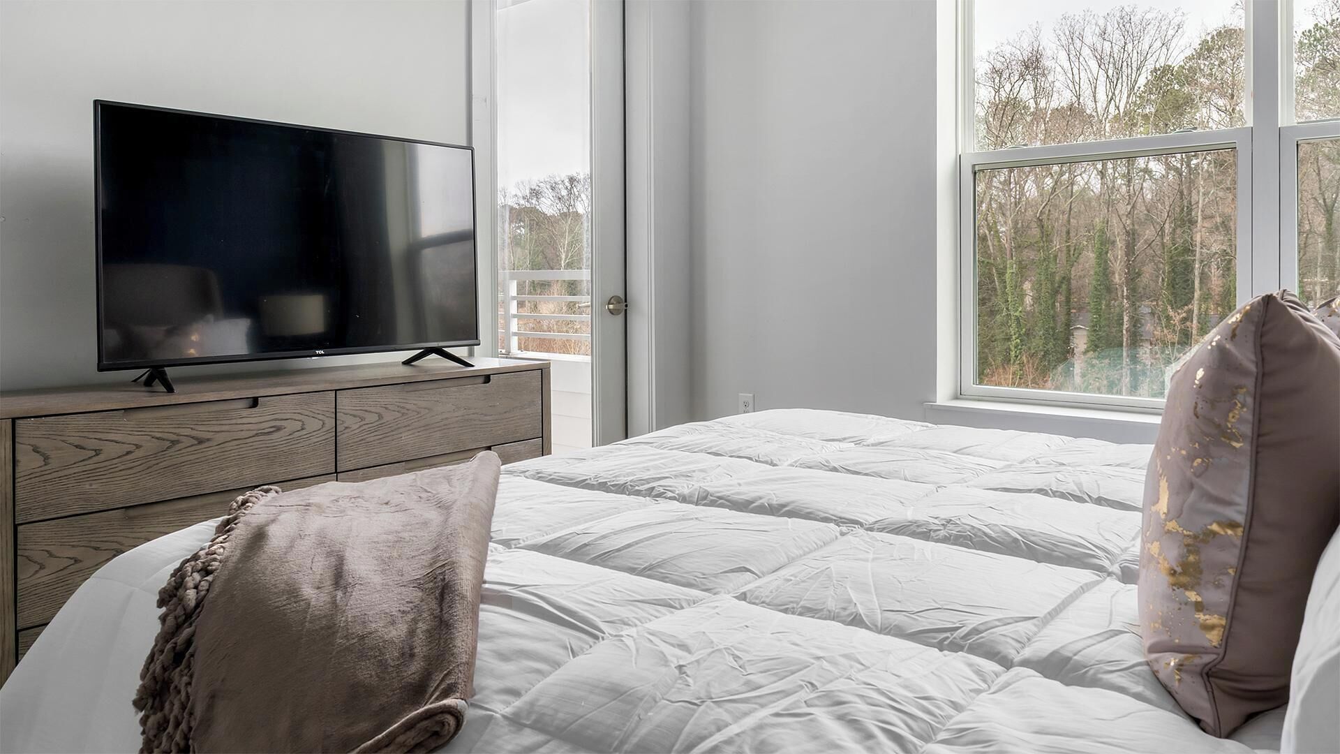 A bedroom with a TV on a wooden dresser, a bed with white bedding, and a window with a view of trees.