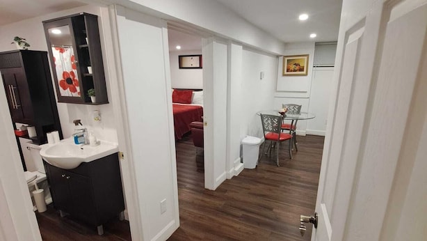 Un couloir mène à une chambre avec une couvre-lit rouge, une salle de bain avec un lavabo blanc et un coin repas avec une table et des chaises.