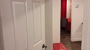 Un couloir avec une porte blanche portant l’inscription « B », qui donne sur une pièce où se trouve un banc rembourré de tissu rouge et un mur orné de motifs.