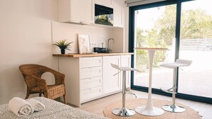 Une cuisine moderne avec des meubles blancs, un coin repas comprenant deux tabourets de bar blancs, un fauteuil en osier et une grande fenêtre donnant sur un paysage verdoyant.