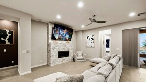 A modern living room with a stone fireplace, a large flat-screen TV, a ceiling fan, and a sectional sofa.