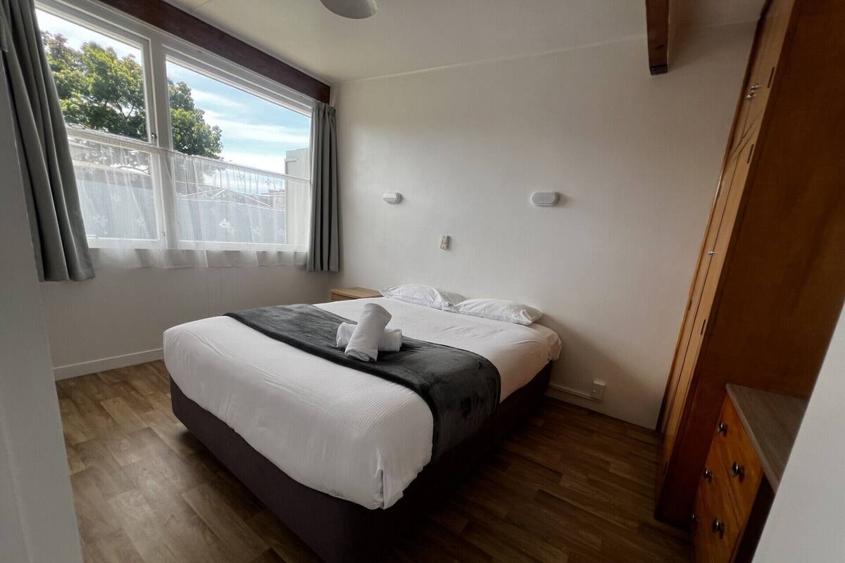 A bedroom with a bed, wooden flooring, a window with curtains, and a wooden wardrobe.