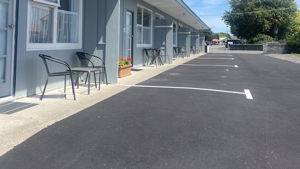 Un motel con una hilera de habitaciones, cada una con ventana y puerta. Se observan sillas exteriores y una jardinera pequeña con flores.