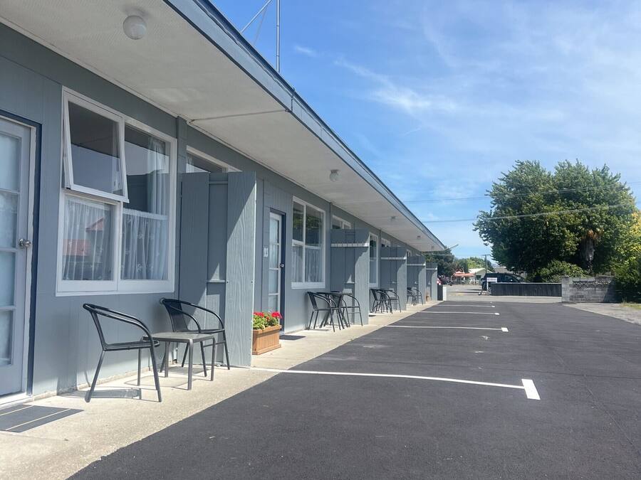 A motel with a row of rooms, each with a window and a door. There are outdoor chairs and a small planter with flowers.