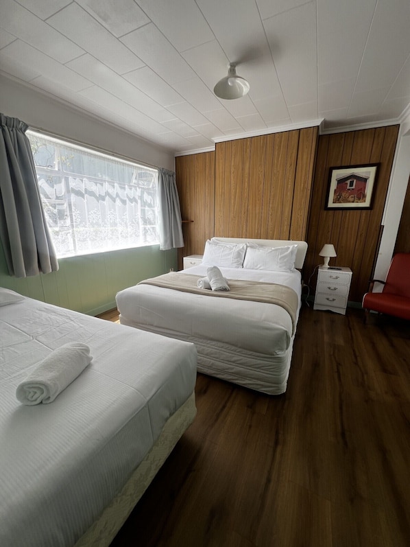 Habitación de hotel con dos camas, un panel de pared de madera, una ventana con cortinas, una mesita de noche con lámpara y una silla roja.