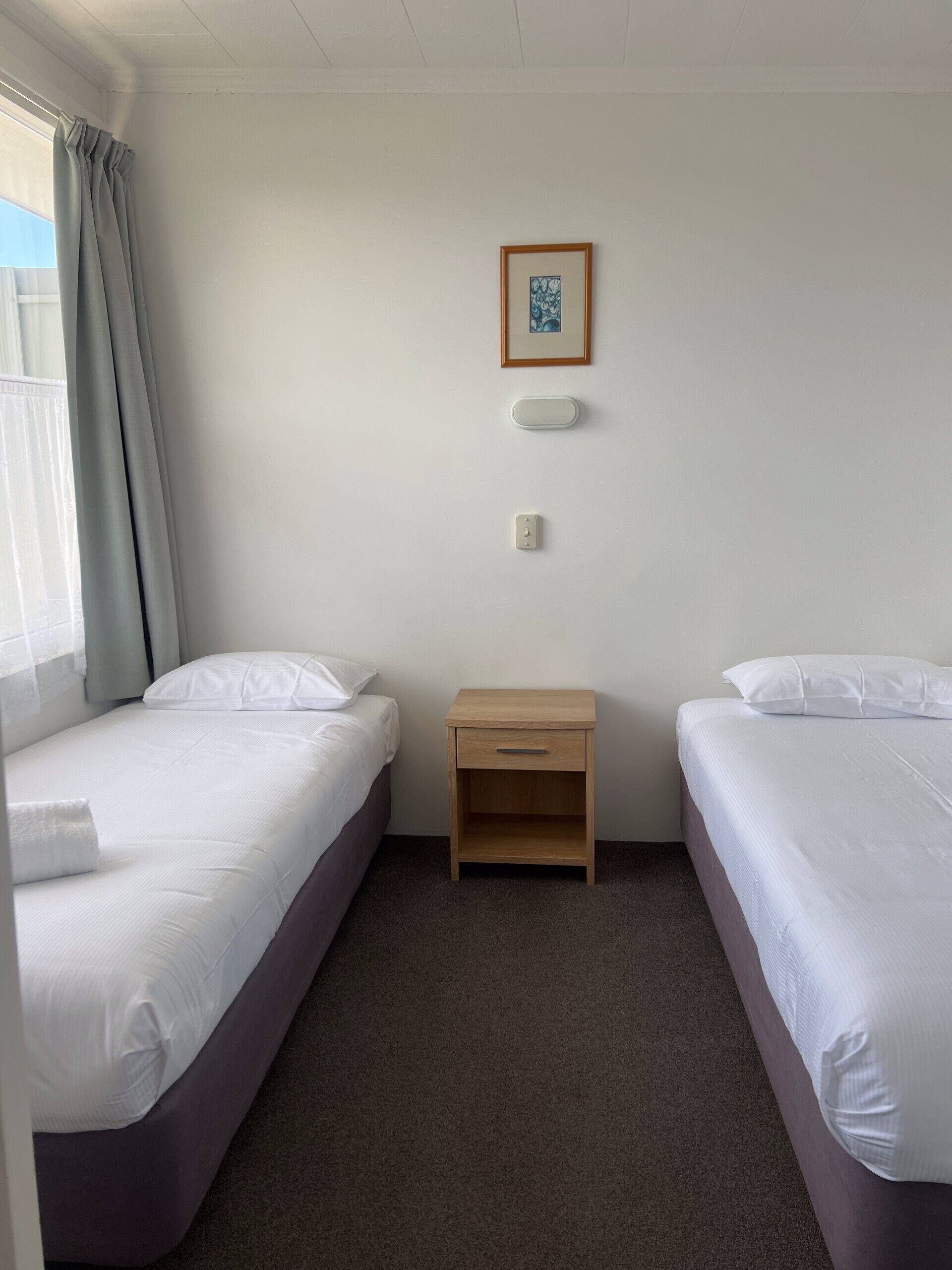 A hotel room with two single beds, a small wooden nightstand, a framed picture on the wall, and a window with curtains.