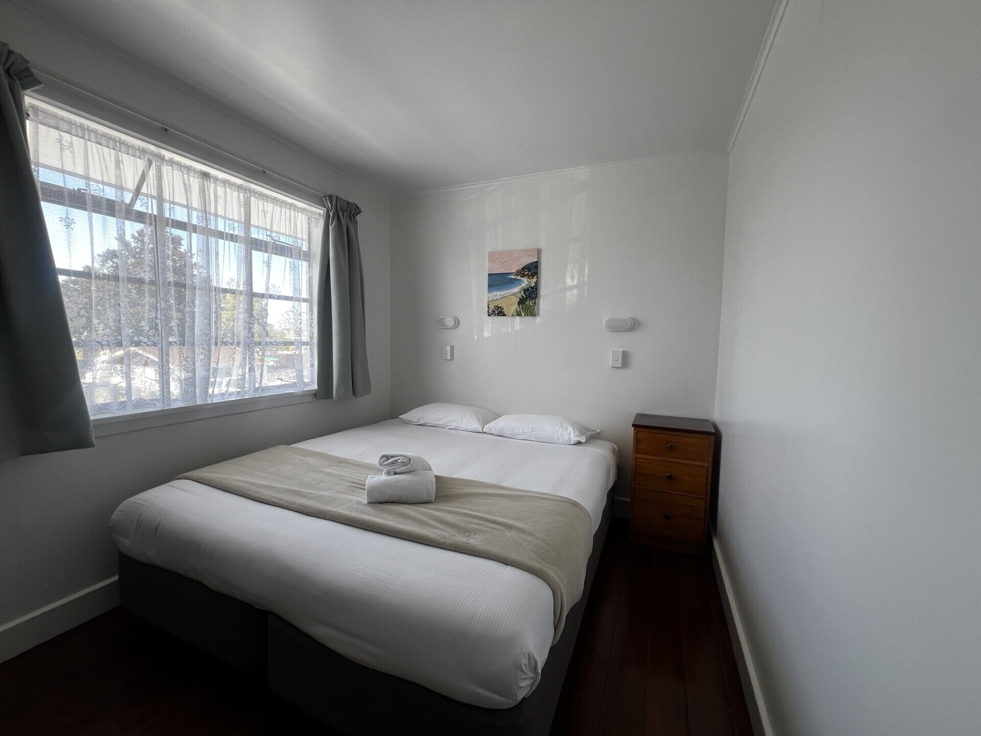Un dormitorio con cama, mesita de noche, ventana con cortinas e imagen colgada en la pared.