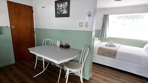 A room with a wooden floor, a white table with two chairs, a bed with a white mattress and pillows, a wooden door, and a wall-mounted light fixture. - Bings Motel (Blenheim)