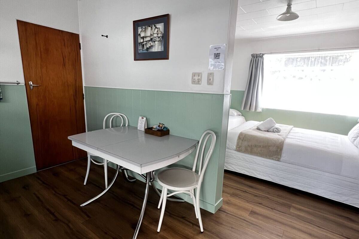 A room with a wooden floor, a white table with two chairs, a bed with a white mattress and pillows, a wooden door, and a wall-mounted light fixture.
