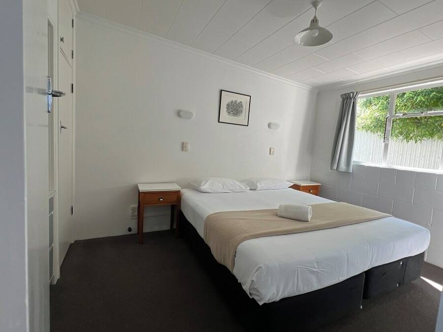 Quarto com cama, mesas de cabeceira, escrivaninha, cadeira, janela com cortinas e luminária no teto.