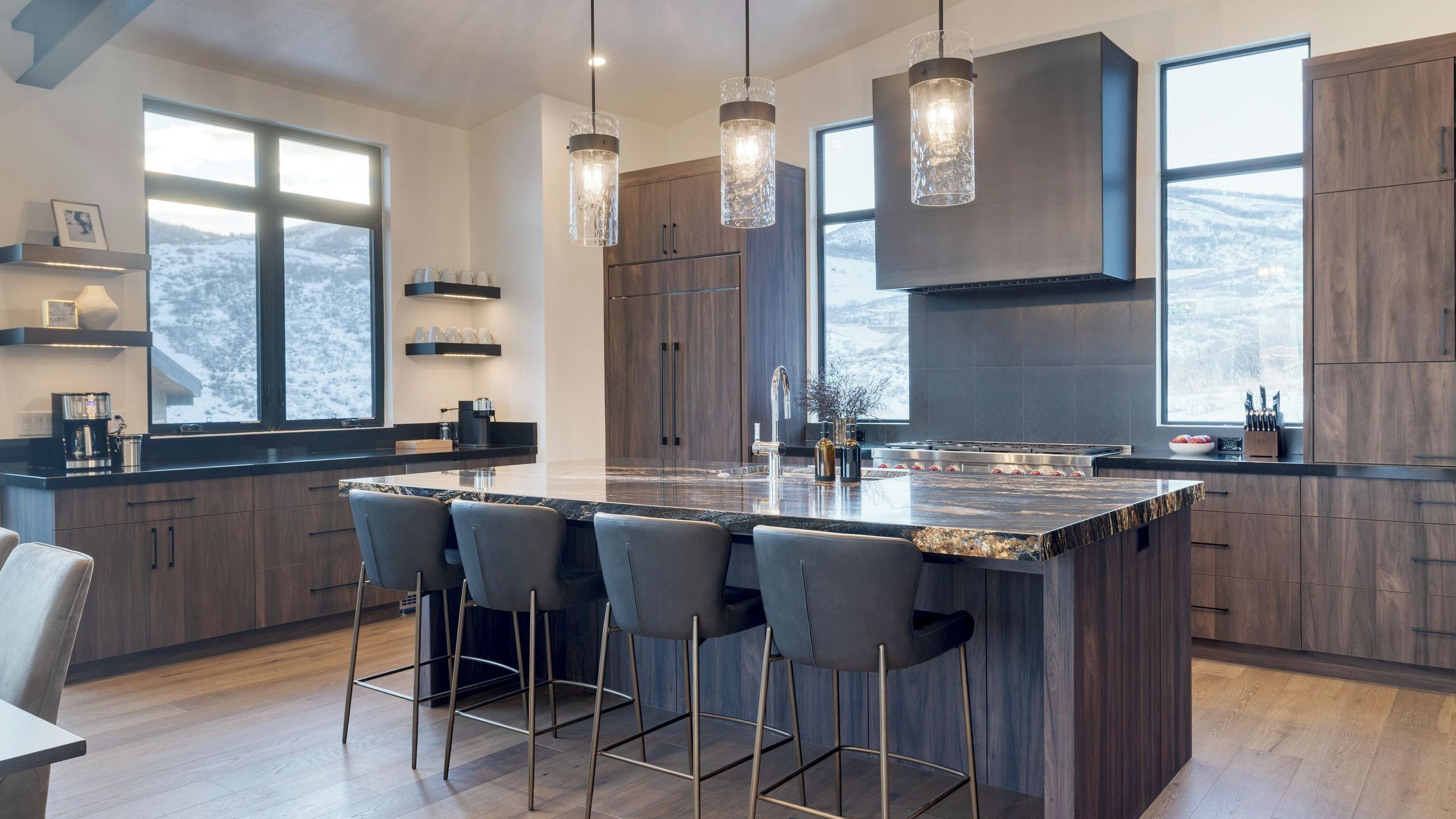 A modern kitchen with a large island, bar stools, pendant lights, and a view of a snowy landscape.