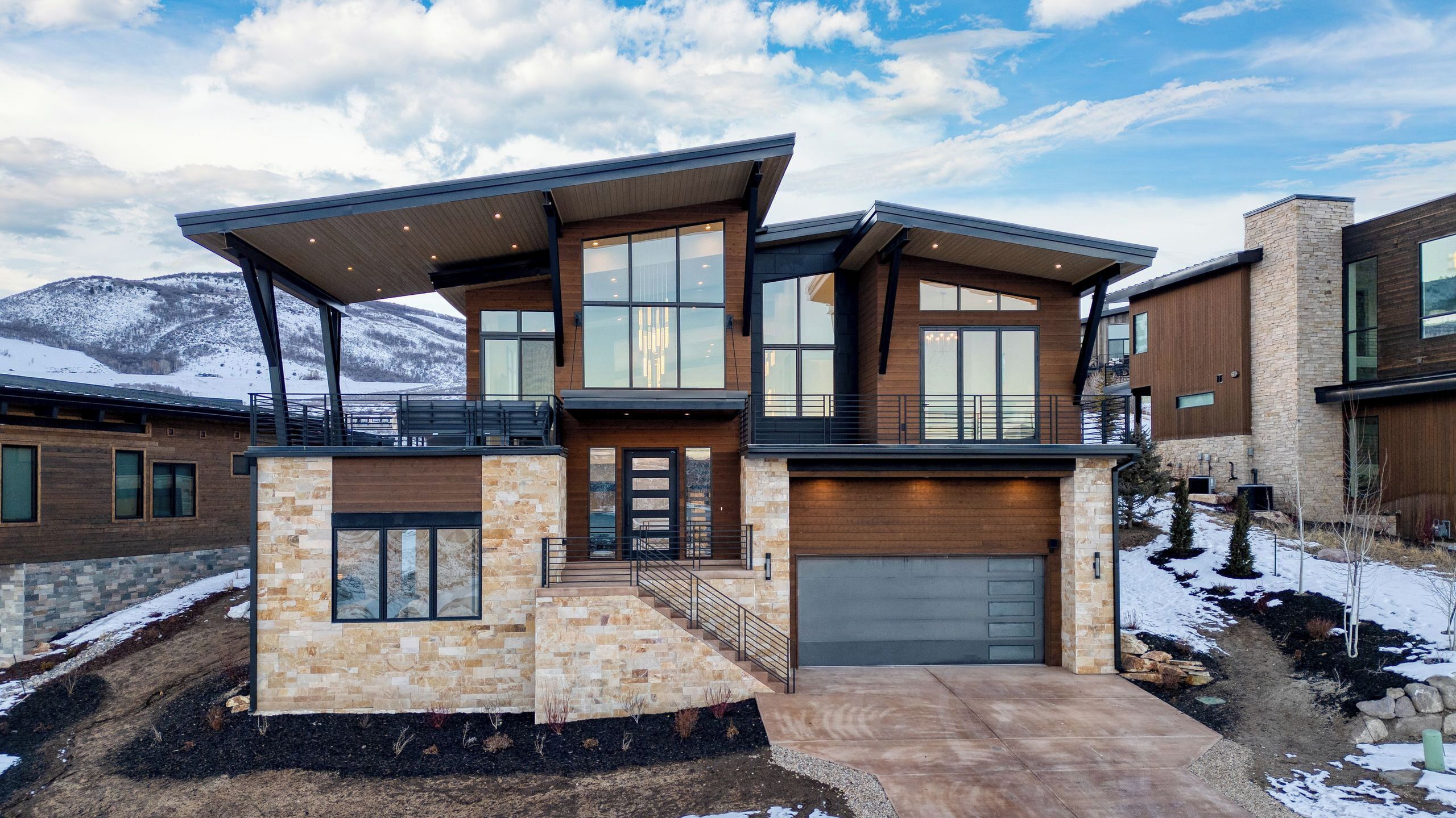 Une maison moderne à deux étages, dotée d’une façade en pierre, de grandes fenêtres et d’un garage.