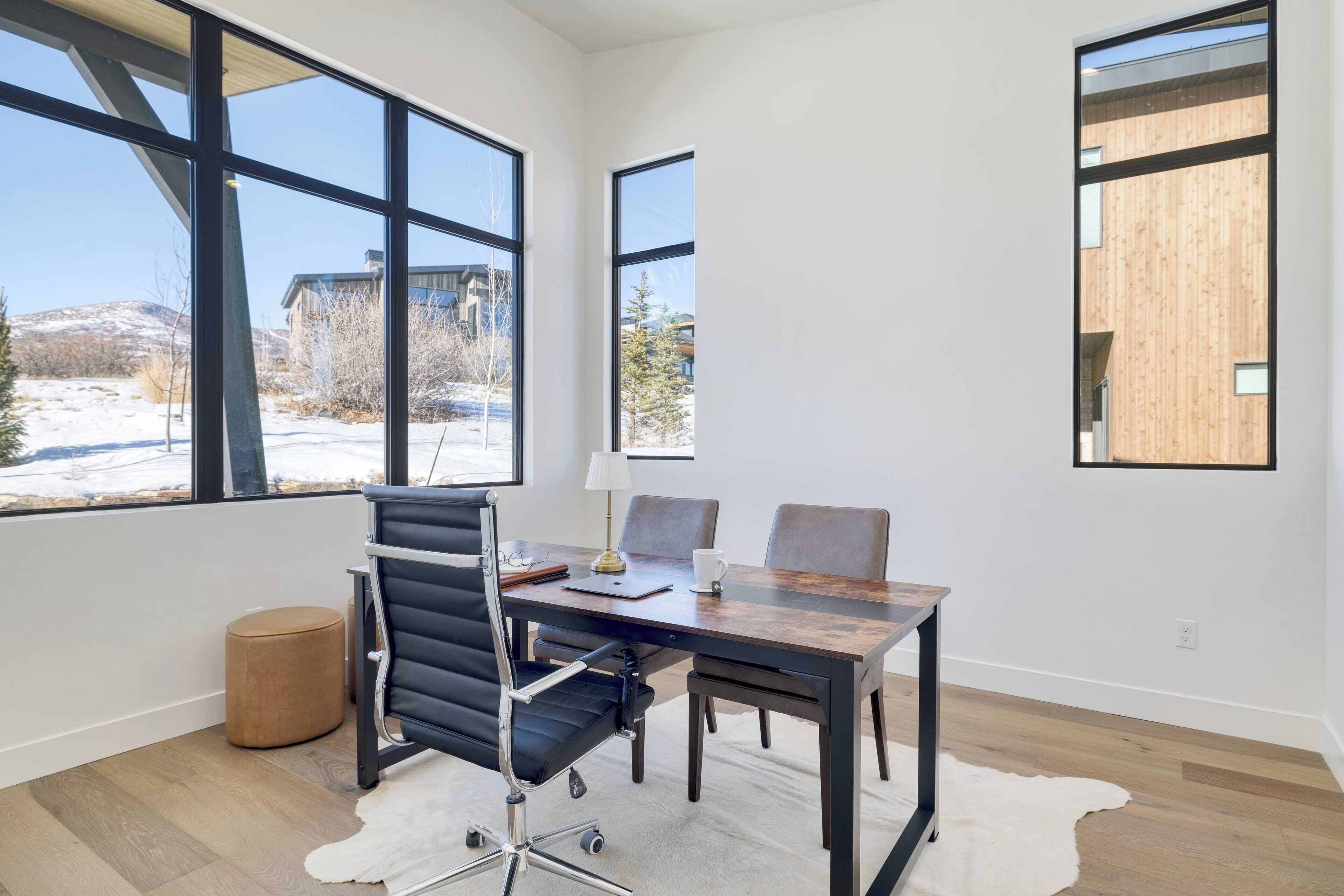 A modern office space with large windows, a leather chair, a wooden table, and a white rug.