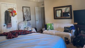 Quarto de hotel com cama, uma cadeira, escrivaninha, ventoinha no teto e espelho.