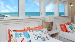 A room with a view of the ocean through large windows, a bed with colorful pillows, a nightstand, and a lamp. - Moonstruck (Port St. Joe)