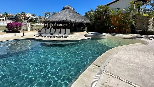 A clear pool with lounge chairs and a thatched-roof structure, surrounded by palm trees and a building. - Condo. Las Quintas, Terraza, Jardín de Paisaje, Zona de Hoteles, Campo de Golf (San José del Cabo)