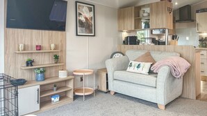 A modern living room with a grey sofa, wooden shelves, a small round table, and a kitchen area with wooden cabinets and appliances. - 2 bedroom accommodation in Ashbourne (Ashbourne)