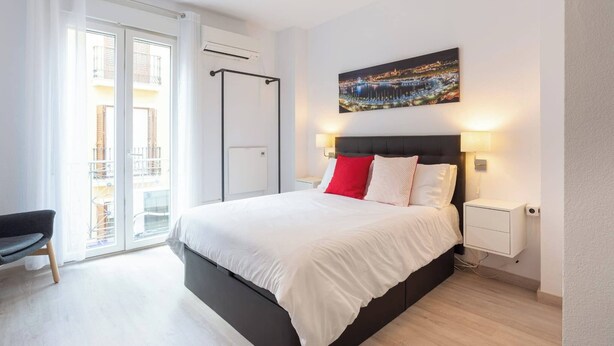Un dormitorio moderno con una cama grande, vistas de la ciudad a través del ventanal y un balcón con barandilla.