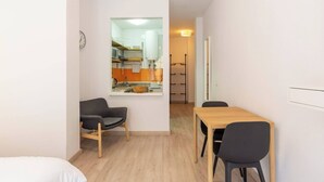 Habitación compacta con cama, una pequeña mesa de comedor con dos sillas y una kitchenette visible a través de una puerta abierta.