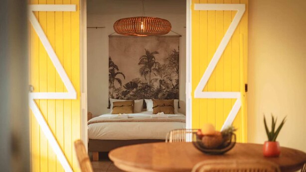 A room with a bed, a dining table, and chairs. There is a large wall tapestry and a hanging light fixture. - Kaz Lorizon (Entre-Deux)