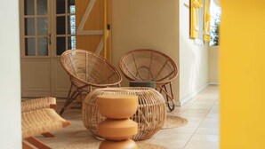 A room with wicker chairs and a stack of stools, a wooden door, and a yellow wall. - Kaz Lorizon (Entre-Deux)
