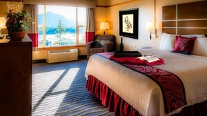 Une chambre d’hôtel avec un grand lit, un bureau agrémenté d’une plante, une chaise, une fenêtre donnant sur les montagnes et une œuvre d’art accrochée au mur.