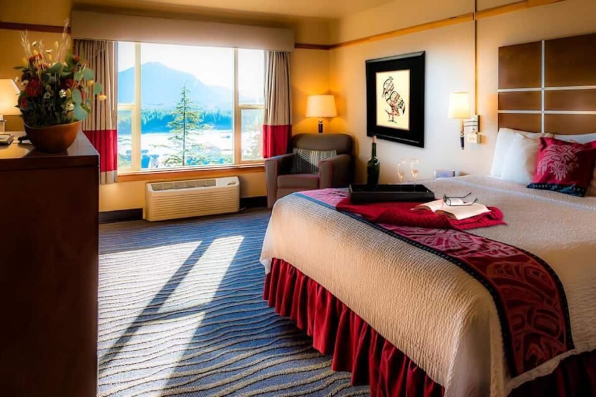 Habitación de hotel con una cama grande, un escritorio con una planta, una silla, una ventana con vistas a la montaña y una obra de arte colgada en la pared.