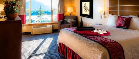 A hotel room with a large bed, a desk with a plant, a chair, a window with a mountain view, and a wall-mounted artwork.