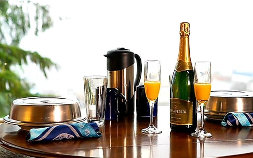 A breakfast setup with a coffee maker, two glasses of orange juice, a bottle of champagne, and a tea set on a wooden table.