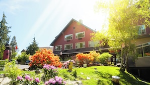 Une maison à deux étages, à l’extérieur rouge, entourée d’un jardin bien entretenu où poussent différentes plantes à fleurs et une pelouse.
