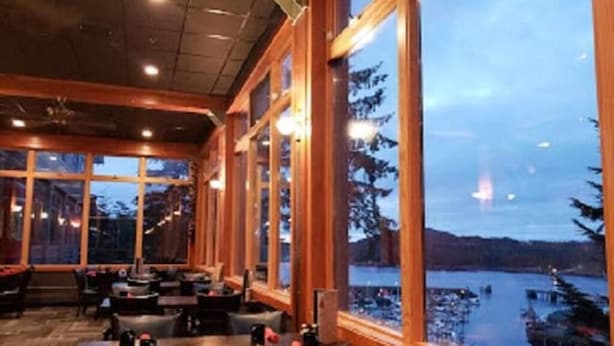 Un restaurant doté de grandes fenêtres donnant sur un plan d’eau, avec un intérieur en bois et des tables accompagnées de chaises.