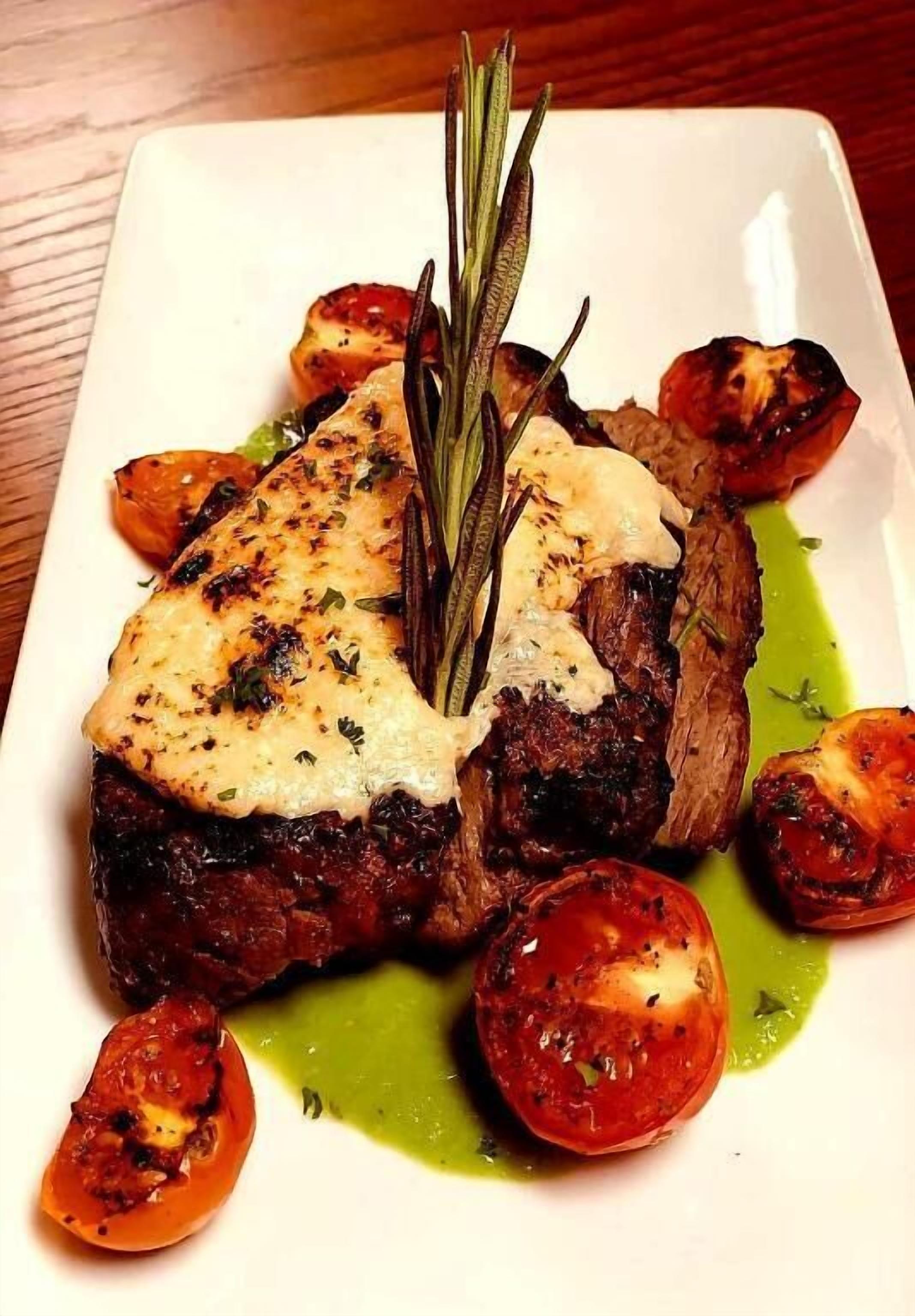 A plate of food with grilled meat, tomatoes, and a sprig of rosemary.