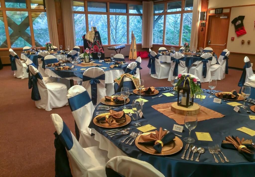 Salão de banquetes com mesas redondas preparadas para um evento formal, com toalhas de mesa azuis e brancas, guardanapos dourados e castanhos e talheres.