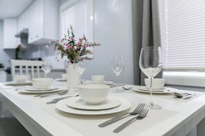 Een net gedekte eettafel met witte borden, kommen, bestek en wijnglazen, met een bloevas op de achtergrond.