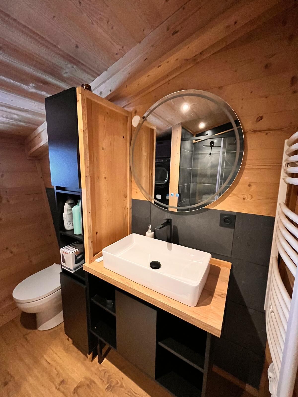 Une petite salle de bain en bois, avec un lavabo blanc, un miroir rond et des toilettes.