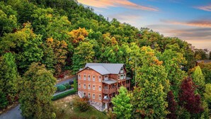 A two-story log cabin with a wrap-around porch, surrounded by dense forest. - A View for All Seasons by Avantstay 12bdr w/ Game Room, Hot Tub, Movie Theater, Deck Views (Sevierville)