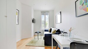A modern, minimalist living room with a sofa, a dining table set for two, and a TV mounted on the wall. - Studio Moderne - 2P- Nanterre (Nanterre)