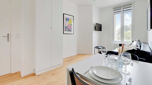 A modern dining area with a white table set for two, a black leather sofa, and a window with blinds. - Studio Moderne - 2P- Nanterre (Nanterre)