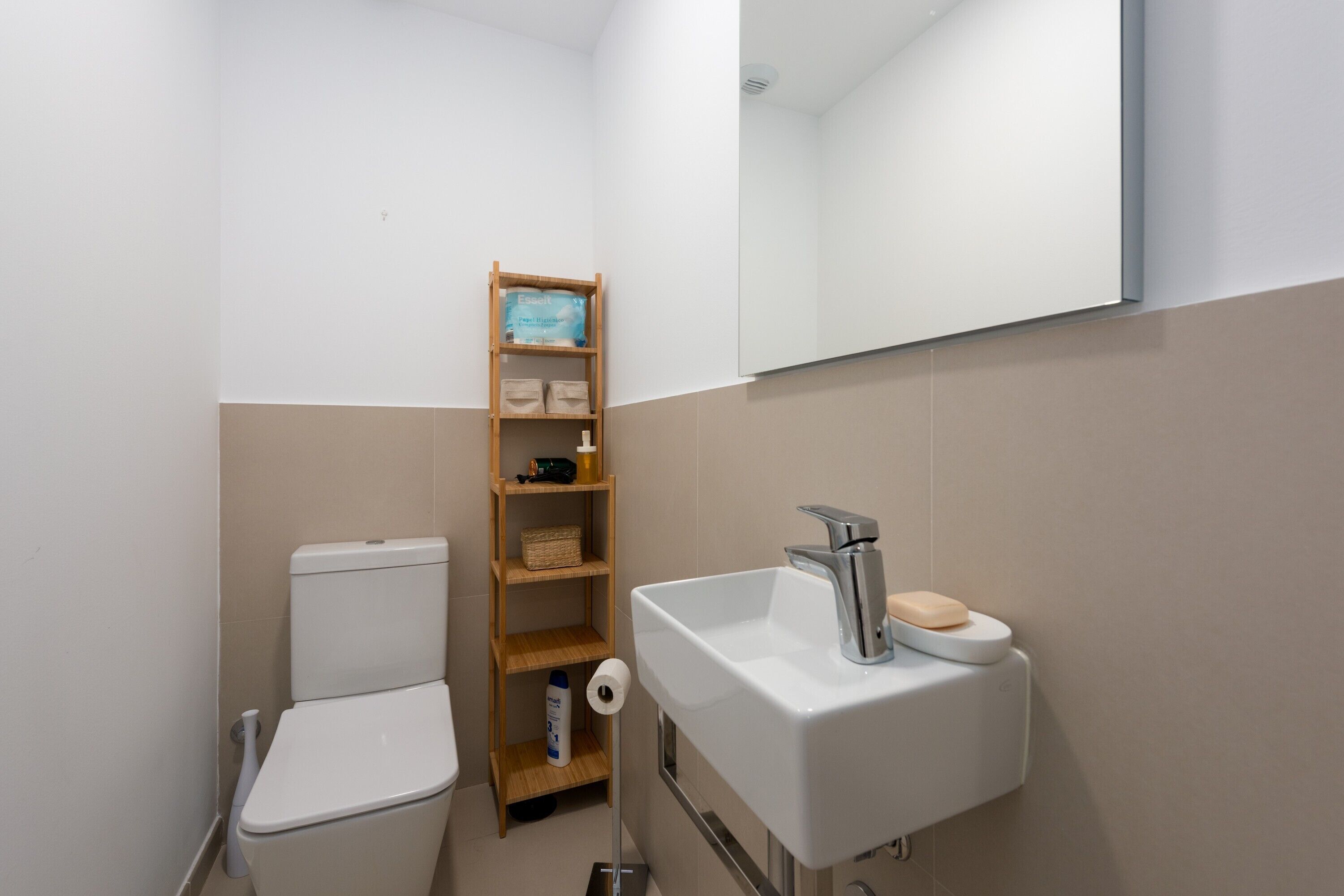 Un baño pequeño con inodoro, lavamanos, espejo y repisa de madera con artículos de aseo.
