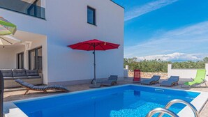 Un área de piscina moderna con una piscina azul cristalina, tumbonas, un paraguas rojo y un edificio blanco con ventanales grandes.