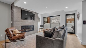 Een moderne woonkamer met een open haard, een bank, een fauteuil en een groot raam met uitzicht op een besneeerd landschap.