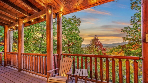 A wooden deck with a wooden chair and table, surrounded by a railing, overlooking a scenic landscape with trees and mountains.